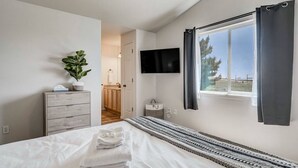 A bedroom with a bed, a TV, a window with curtains, a plant, and a view of the outdoors.