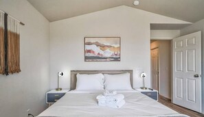 A hotel room with a bed, a painting on the wall, a nightstand, a lamp, and a door.
