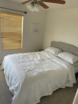 Een slaapkamer met een bed, een plafondventilator, een raam met zonwering en een aan de muur bevestigd fotolijstje.