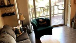 Un salon avec un canapé, un fauteuil vert et une table basse en bois. Une lampe est posée sur une table d’appoint, et des verres à vin se trouvent sur une étagère.