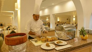 A chef preparing food in a well-lit kitchen with a buffet setup, including a large metal container and a terracotta dish. - HOTEL DAR TAOUFIK (Djerba Midun)