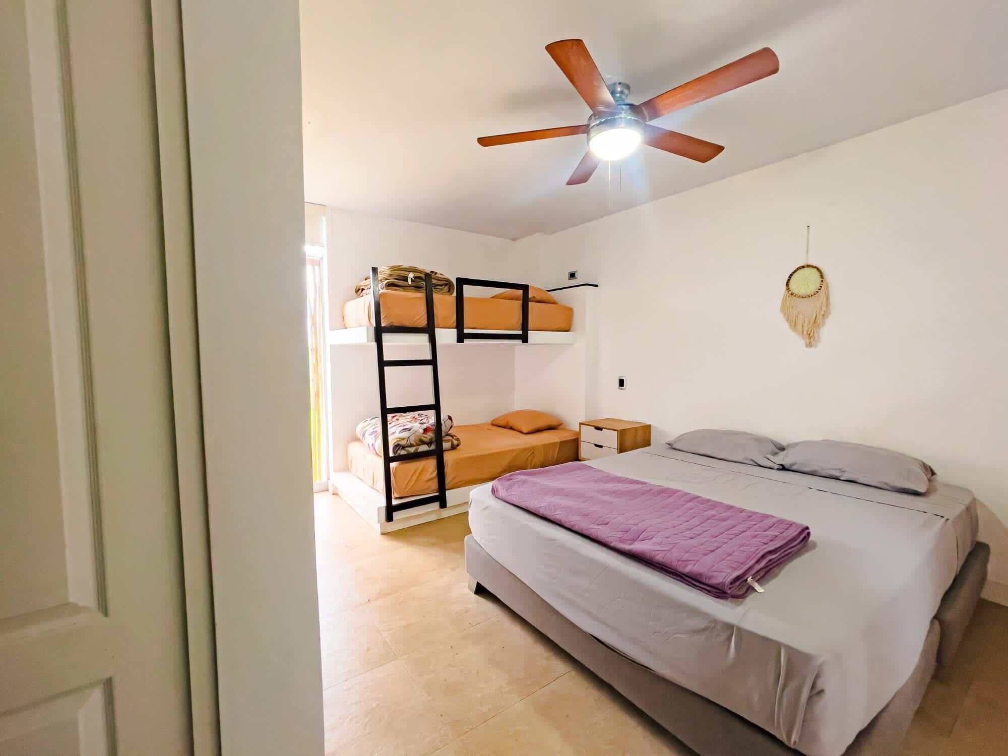 Ein Schlafsaalzimmer mit Etagenbetten, einem Bett, einem Deckenventilator und einem Traumfänger an der Wand.