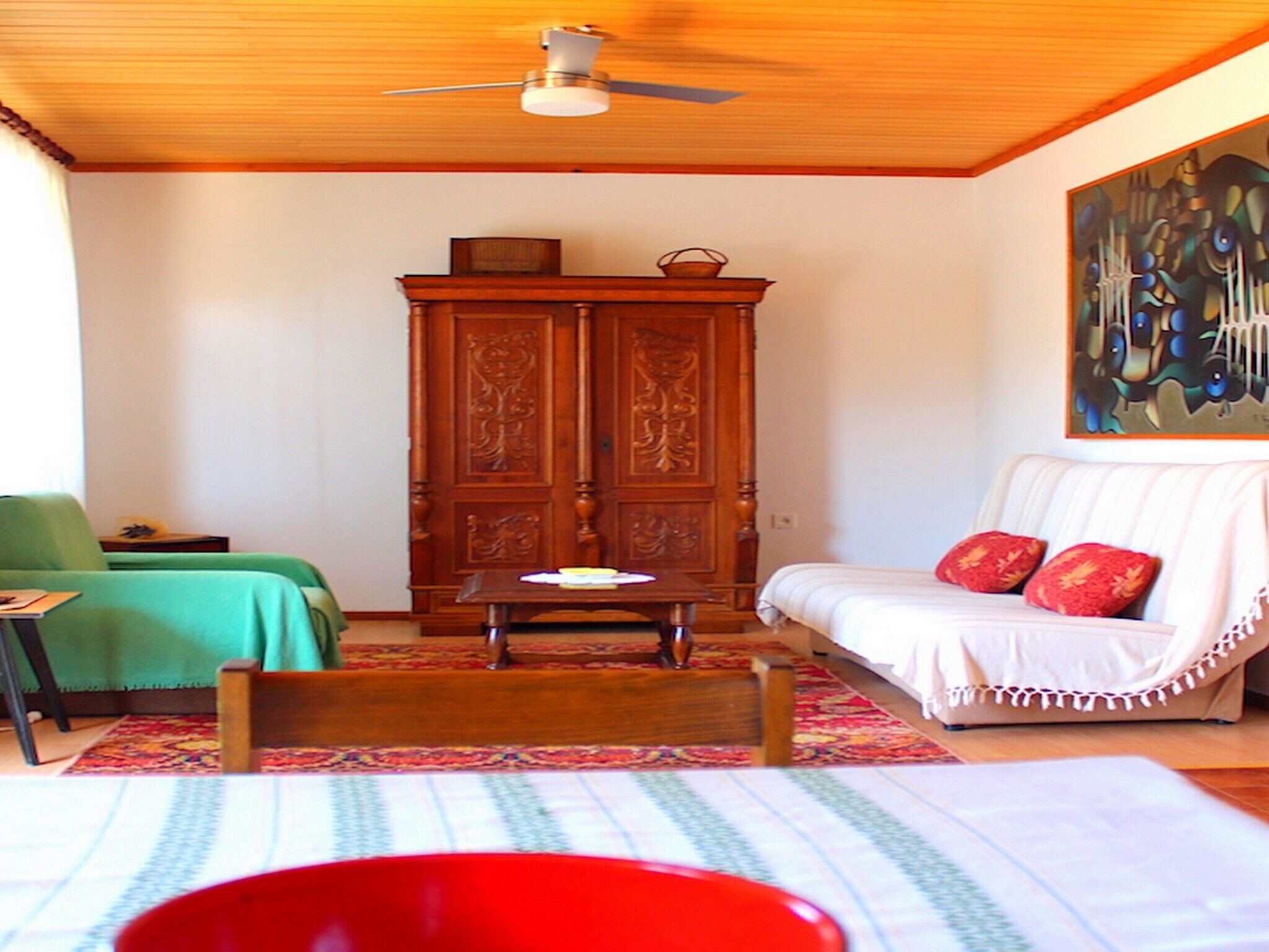 A room with a wooden ceiling, a green chair, a white sofa with red cushions, a wooden cabinet, and a painting on the wall.