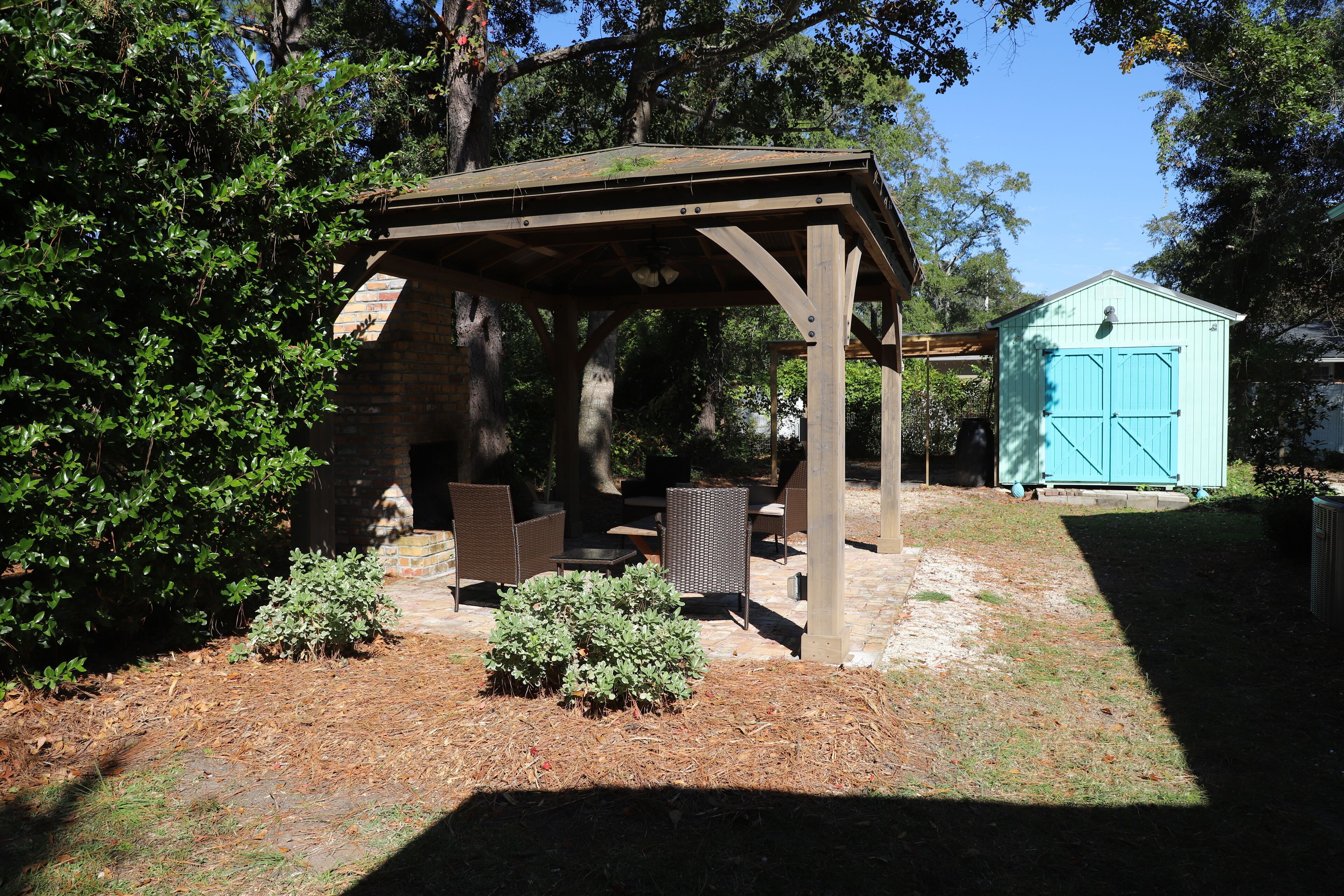 A wooden gazebo with a bench and table, surrounded by trees and a small shed.