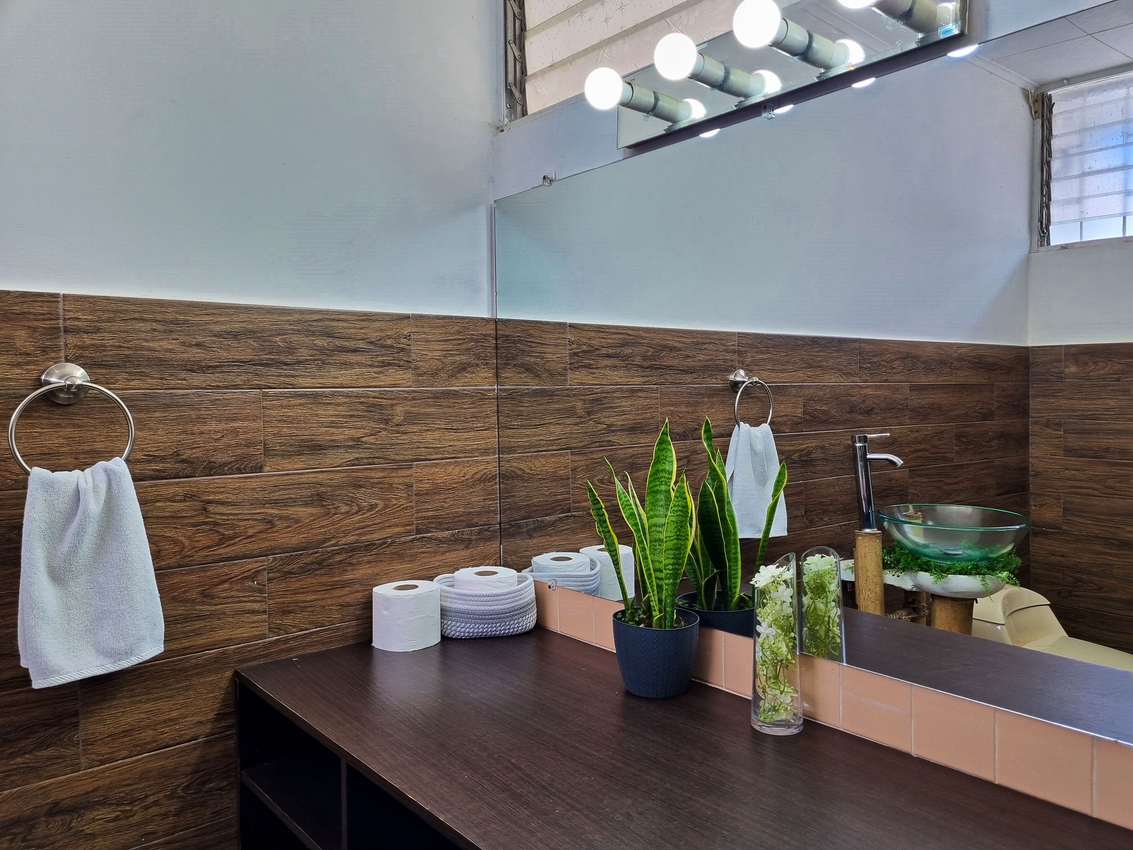 Un baño moderno con una pared de madera, un espejo grande, un lavabo con un recipiente de vidrio y diversos artículos de tocador en la encimera.
