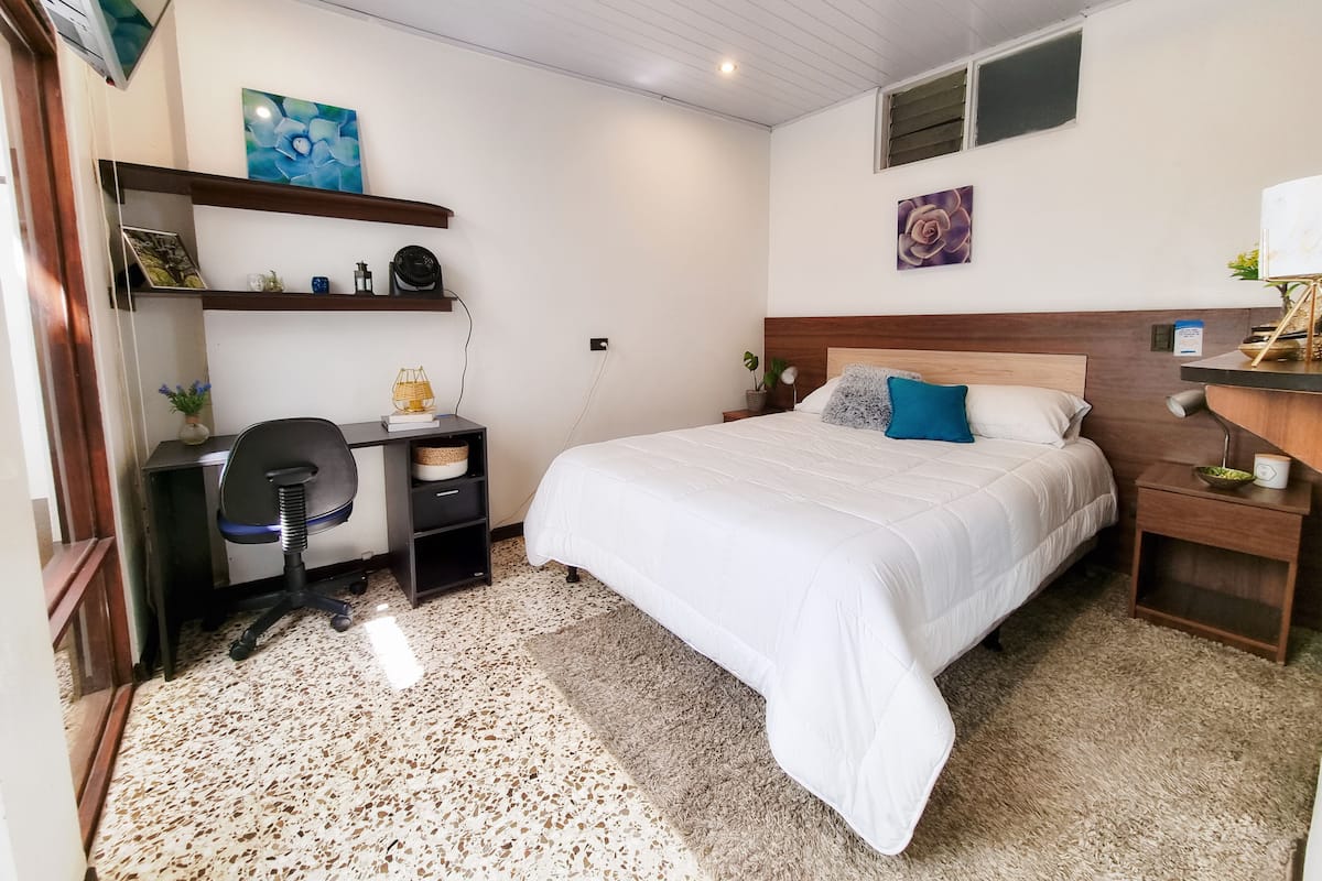 A bedroom with a bed, a desk, and a chair. There is a small table with a lamp and a plant. The room has a tiled floor and a ceiling with recessed lighting.