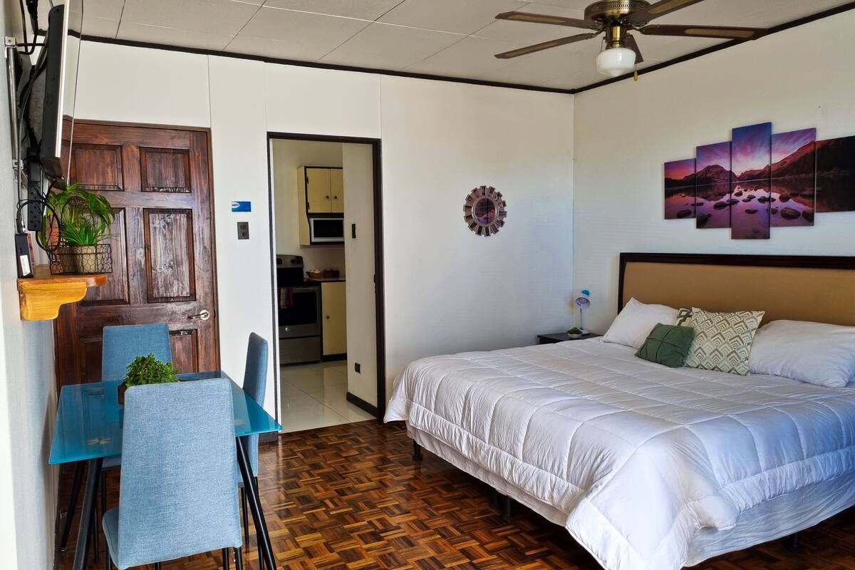 A bedroom with a bed, a desk with chairs, a ceiling fan, and a kitchen in the background.