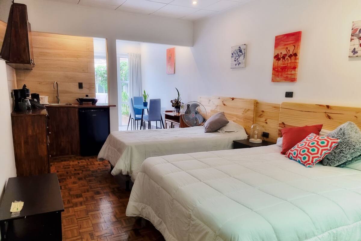 A hotel room with two beds, a wooden headboard, a small table, a desk, a chair, and a window with curtains.