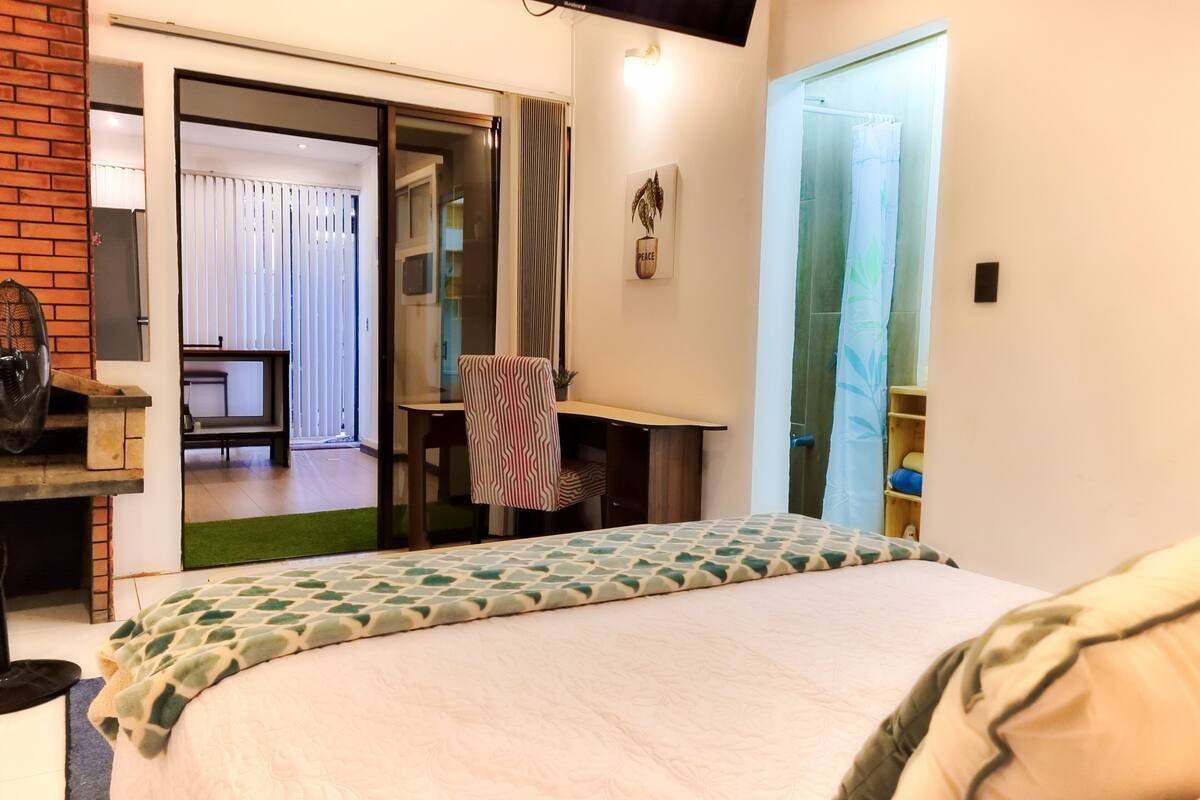 A bedroom with a bed, a desk, a chair, a window with curtains, and a door leading to another room.