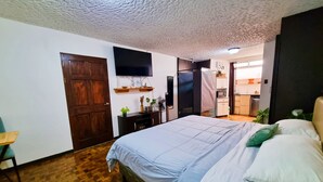 A bedroom with a bed, wooden floor, a TV mounted on the wall, a door, and a small table.