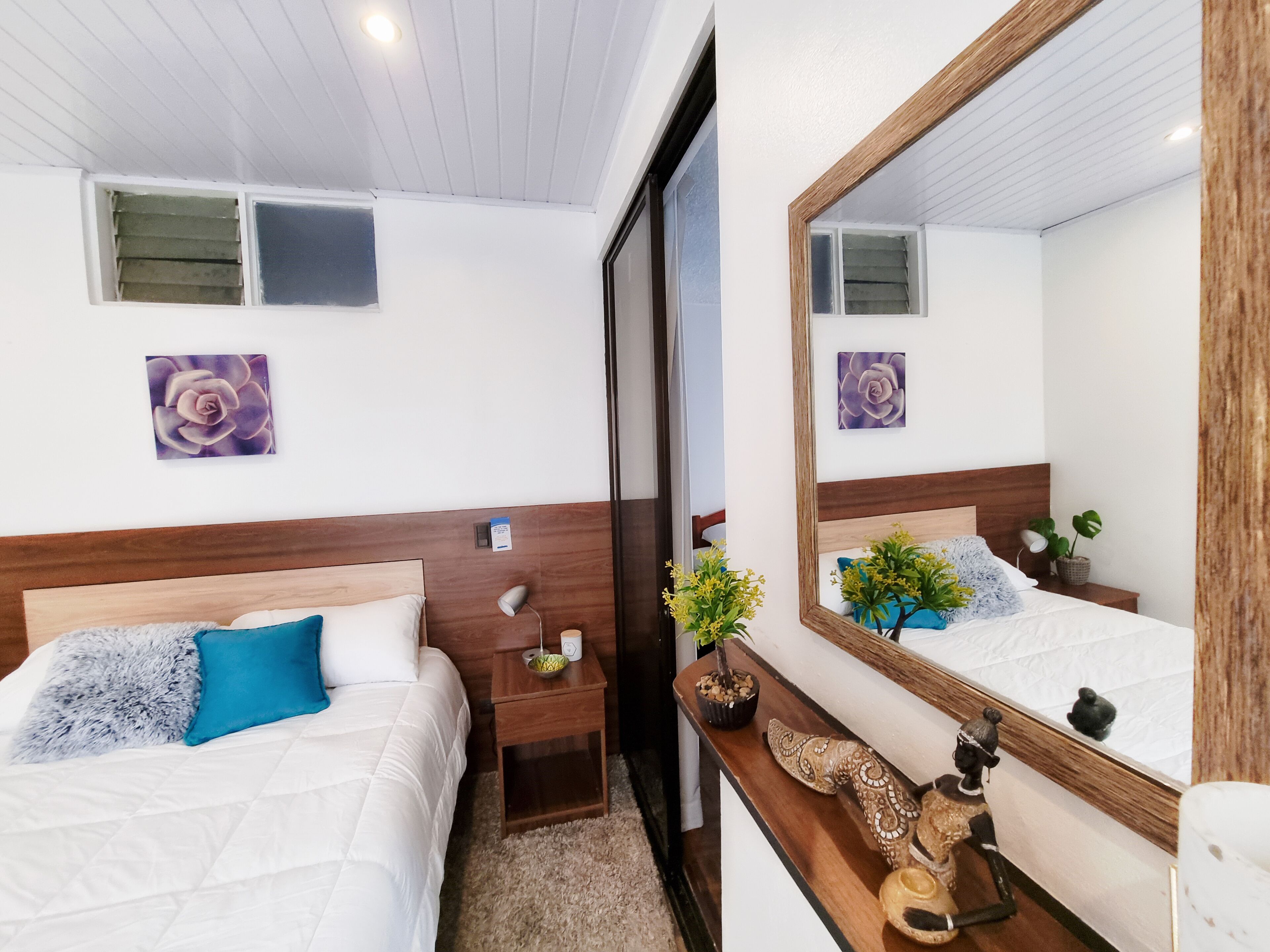 Habitació d'hotel amb dos llits, un amb capçalera de fusta, una taula de nit amb una làmpada, una planteta i un mirall.