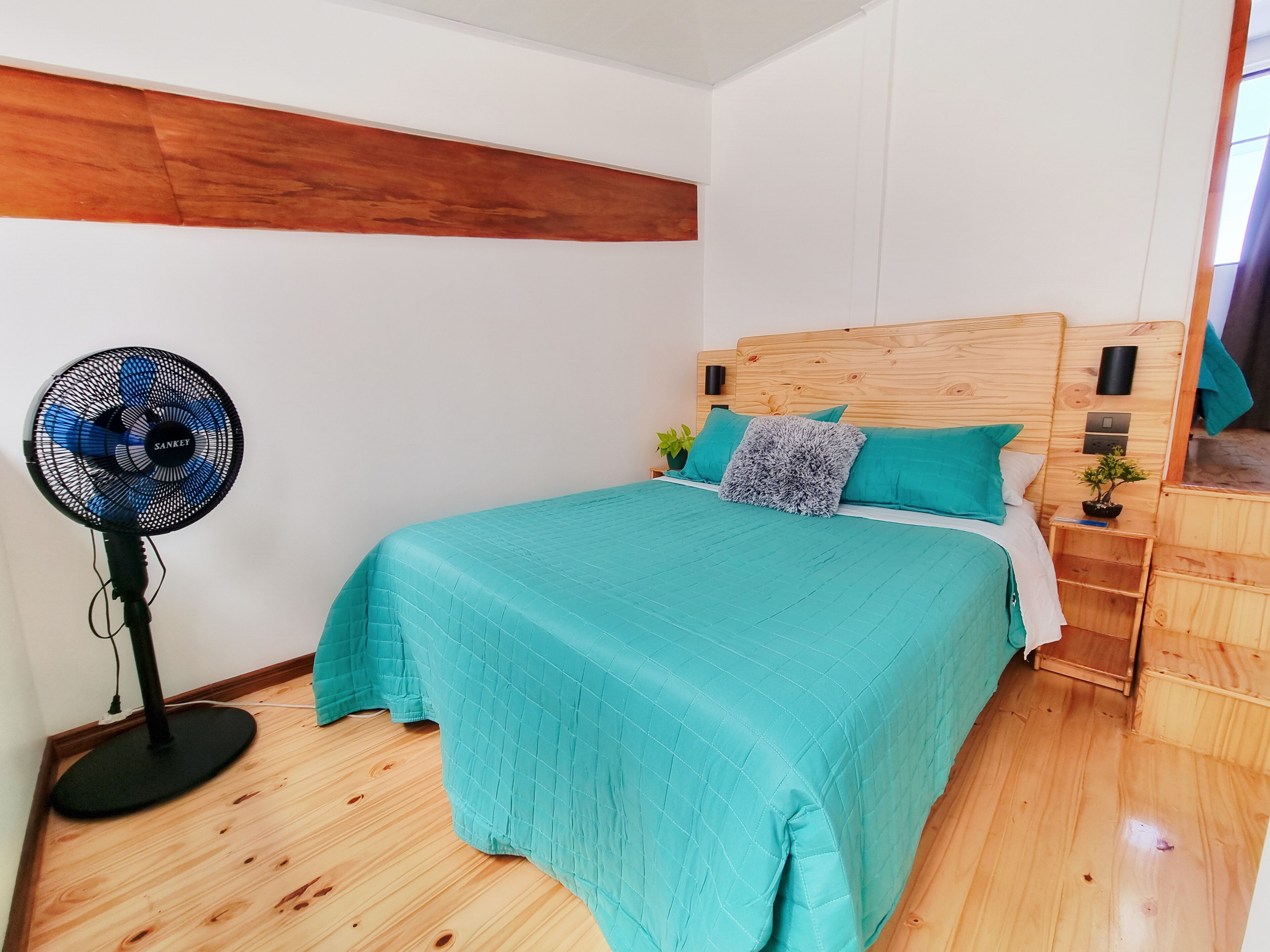 Een slaapkamer met een bed, een staande ventilator en een houten hoofdbord.