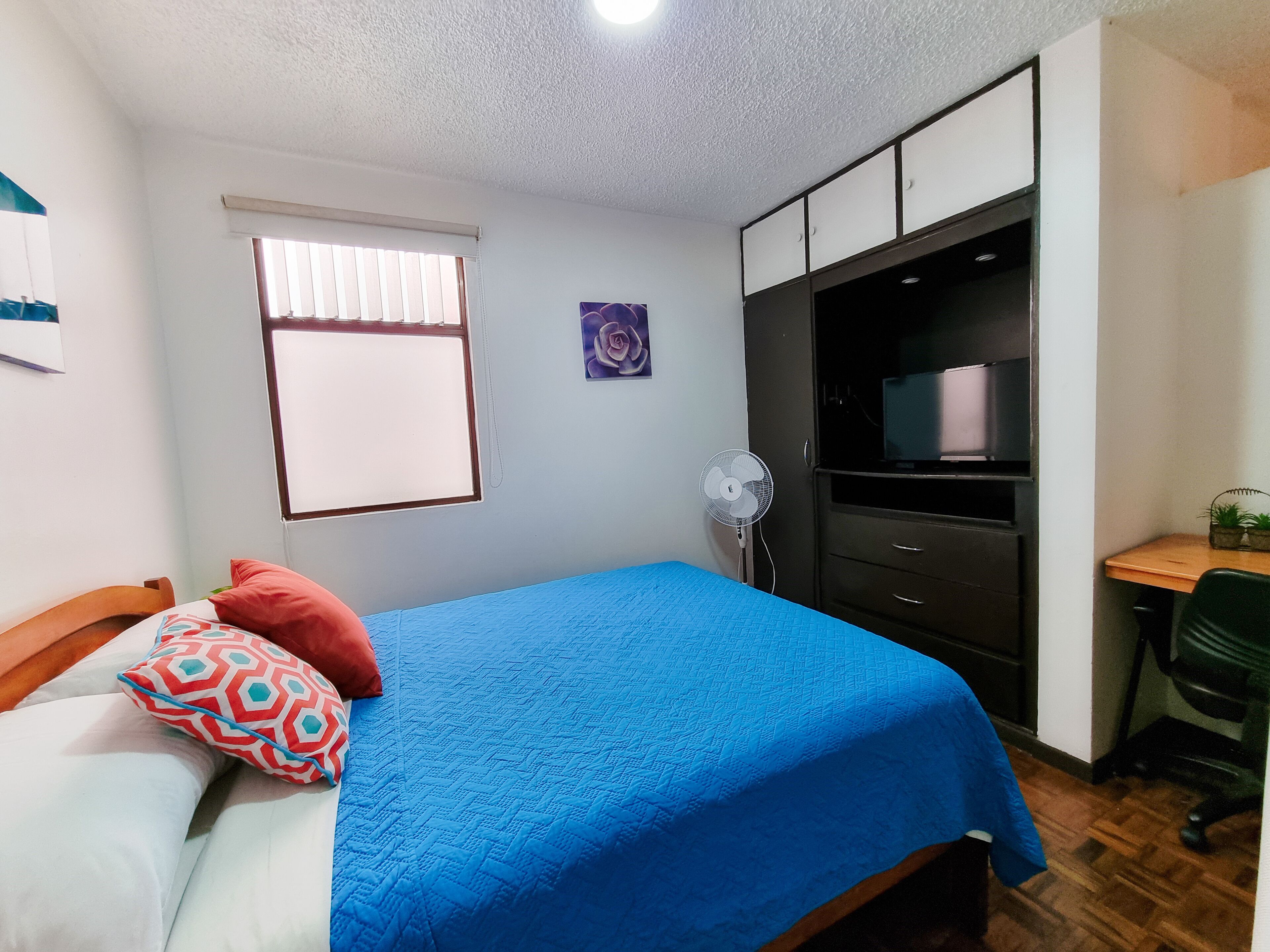 A bedroom with a bed, a desk, a TV, and a window.