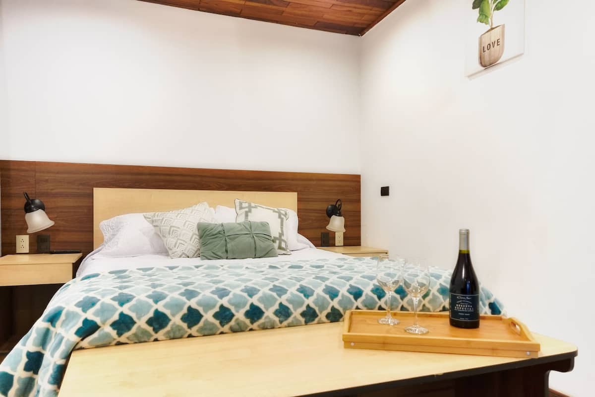 A neatly made bed with patterned bedding, a bedside table with a bottle and glasses, and a wall-mounted sign.