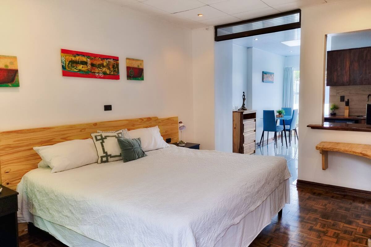 A bedroom with a wooden headboard, a large bed with white bedding, a desk with a chair, and a kitchenette area.