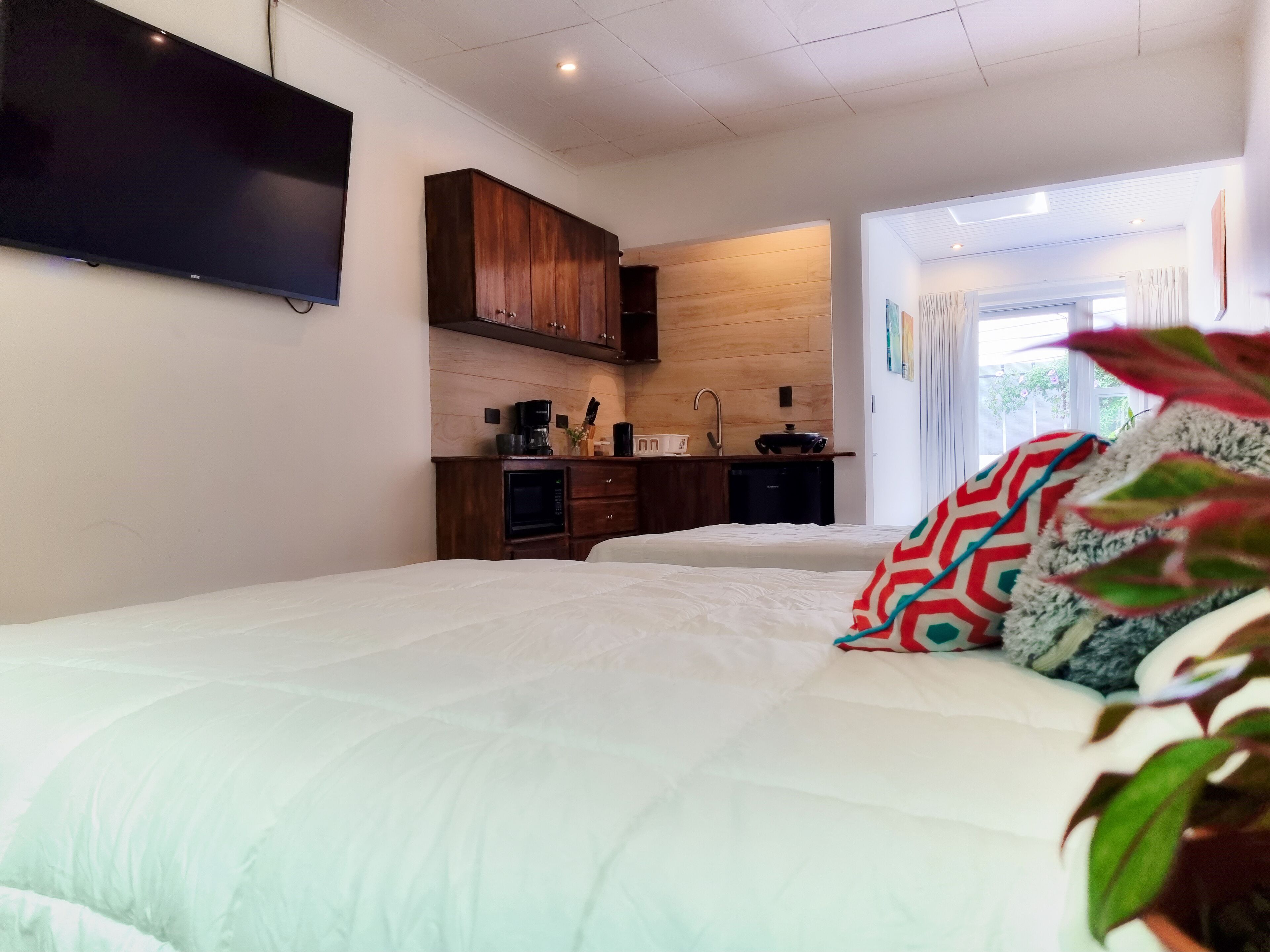 A bedroom with a large bed, a TV mounted on the wall, wooden cabinets, a kitchenette, and a window with curtains.
