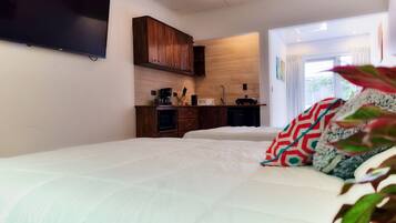 A bedroom with a large bed, a TV mounted on the wall, wooden cabinets, a kitchenette, and a window with curtains.