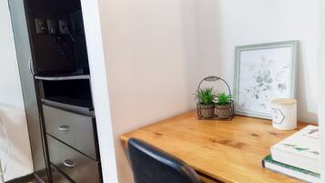 Un rincón de una habitación con un escritorio de madera, una silla negra, una maceta pequeña con planta, un cuadro enmarcado y una taza blanca.