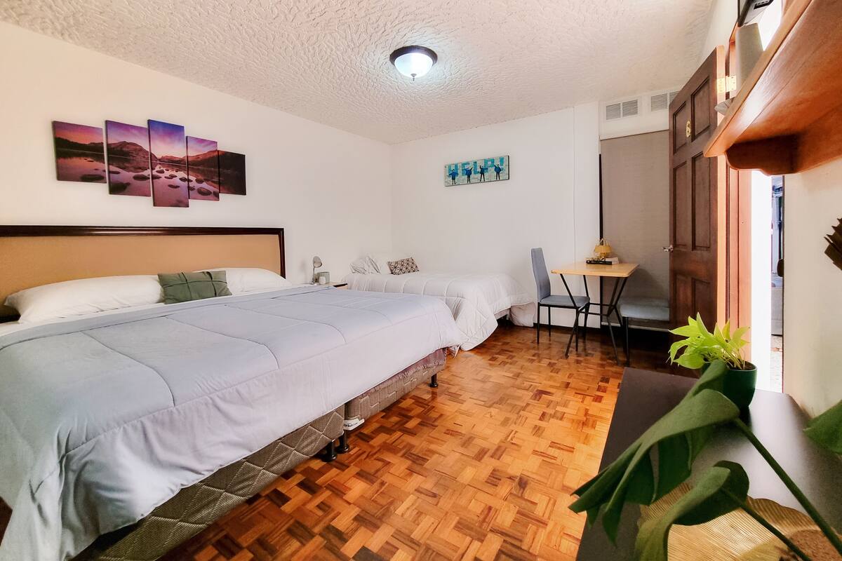 A bedroom with a large bed, a smaller bed, a desk with a chair, a ceiling light, and a wall with artwork.