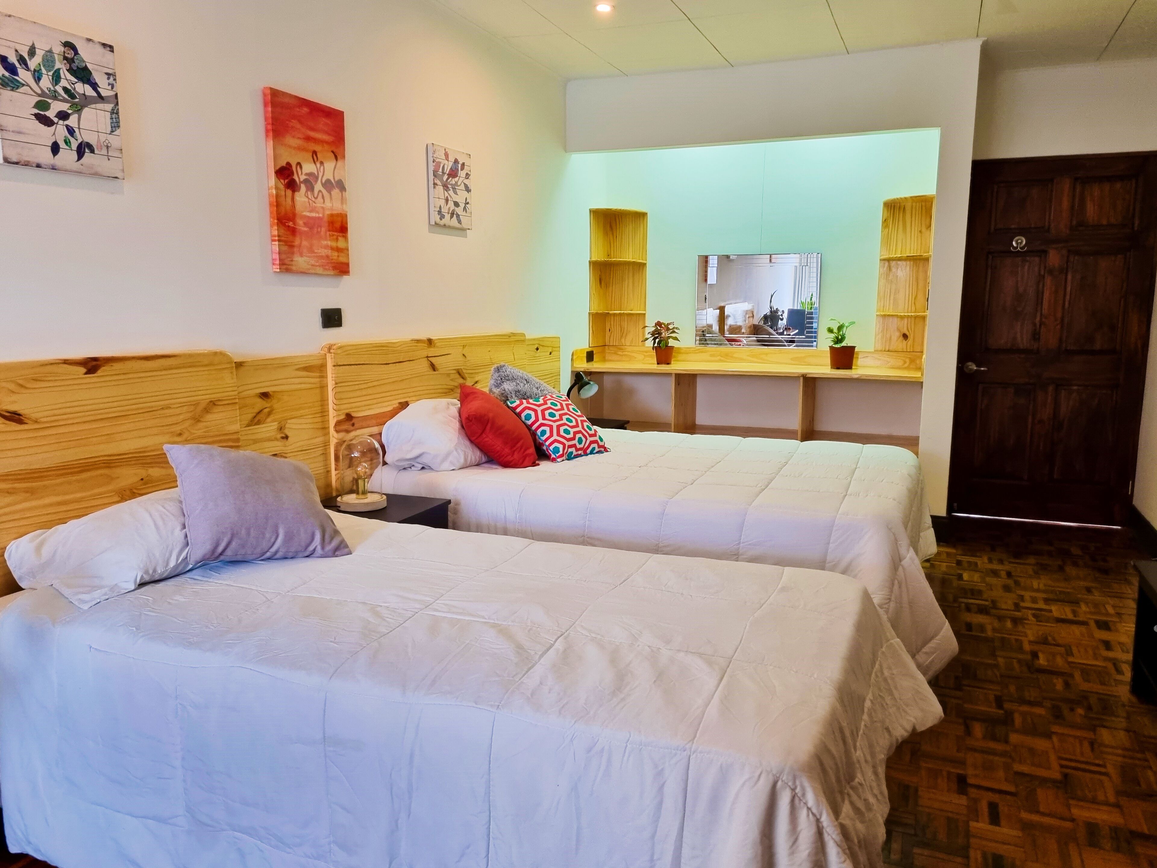 Une chambre avec deux lits, des têtes de lit en bois, un miroir au-dessus d’un bureau en bois et une porte en bois.