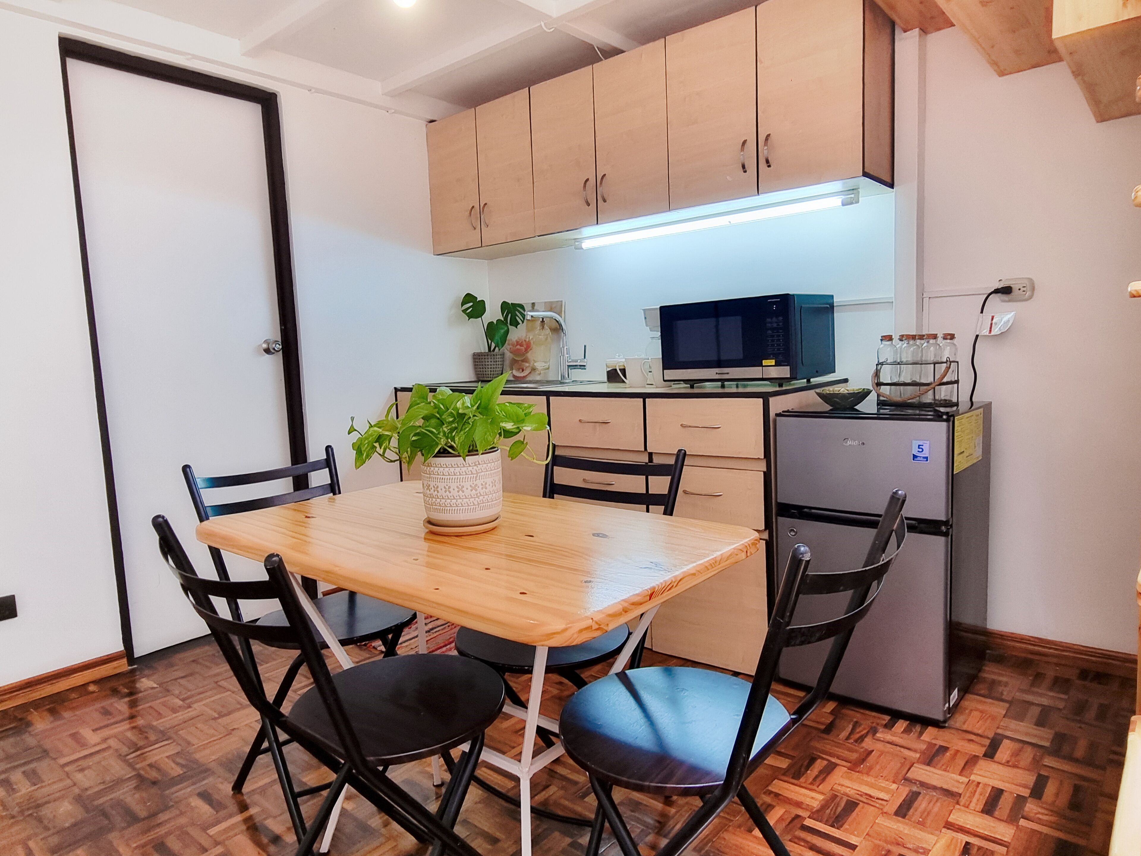 A compact kitchen with wooden cabinets, a small refrigerator, a microwave, and a dining table with chairs.