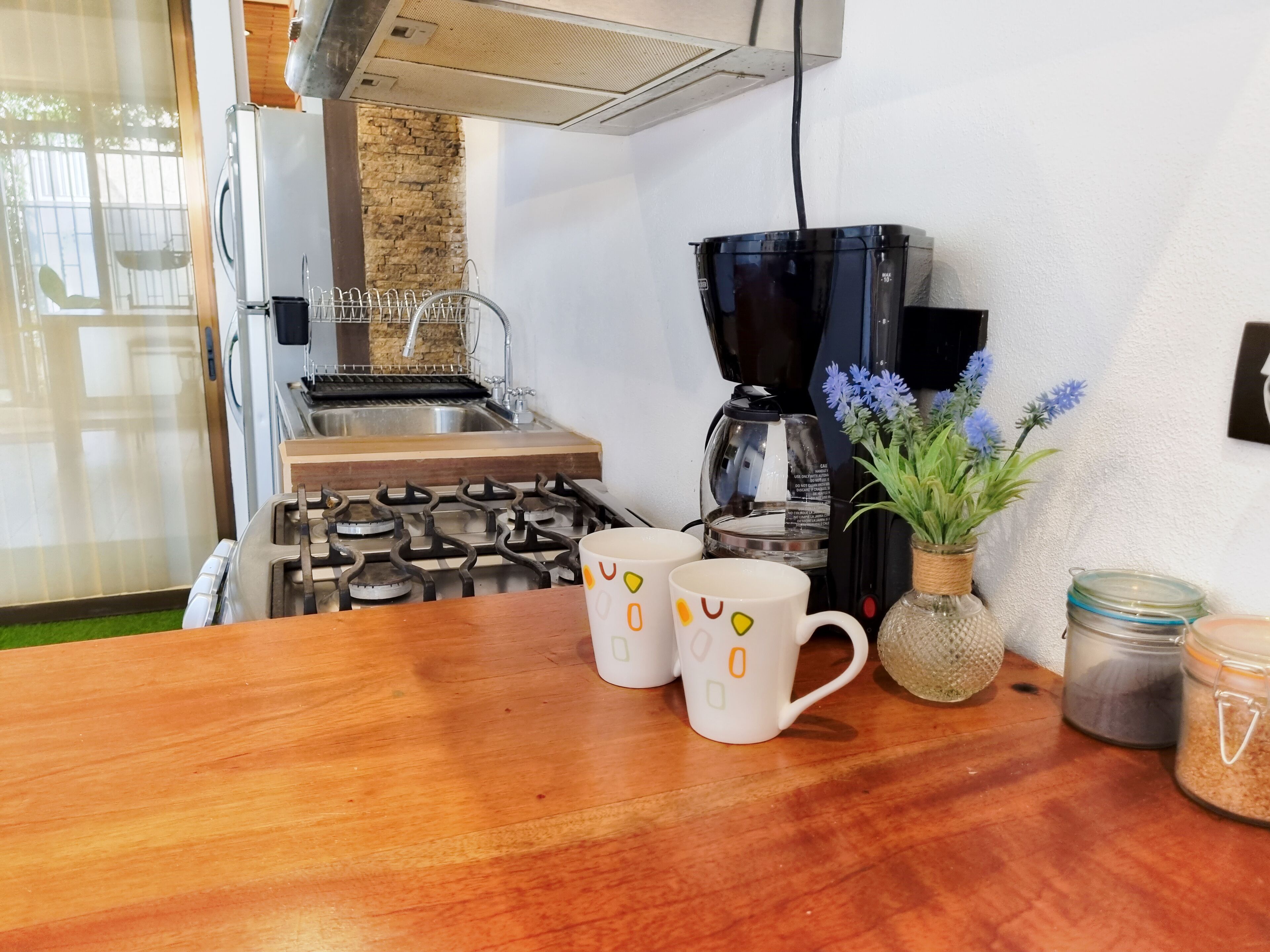 Una cocina con estufa a gas, cafetera y una mesa de madera con tazas y un jarrón con flores.