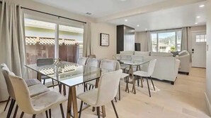 A spacious living area with a glass dining table surrounded by chairs, a sofa, and a large window offering a view of the outdoors. - Spacious Pacifica Beach Home  Sleeps 14 Game Room (Pacifica)