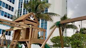 A wooden playground with a slide, swings, and a climbing structure, set against a backdrop of a modern building and palm trees. - 2bd2bth Partial Ocean view Ste w/ balcony, Pool, nearby dining, beach access (Miami Beach)