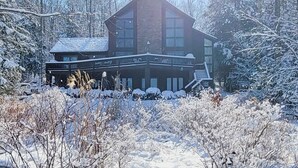 A two-story lodge with a large deck and a covered porch, surrounded by snow-covered trees and a clear sky.