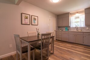 Un coin repas avec une table et des chaises en bois, une cuisine avec des meubles gris et une fenêtre, et deux plantes en pot encadrées accrochées au mur.