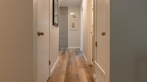 A hallway with wooden flooring, white walls, and a door leading to a room with a white ceiling and recessed lighting.
