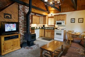 A cozy living room with a fireplace, wooden furniture, a television, and a kitchen area with wooden cabinets and appliances. - Serene retreat is just steps to the National Forest (Big Bear)