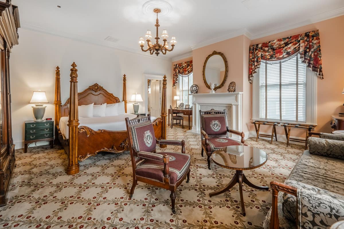 A classic hotel room with a large bed, two armchairs, a small table, a fireplace, and a large window with curtains.