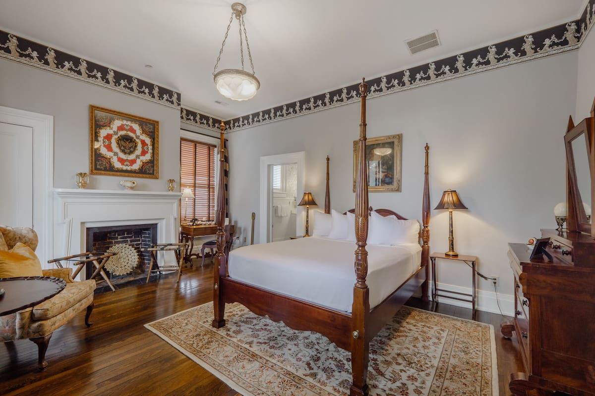 A four-poster bed with a white bedspread, a fireplace, a chandelier, and a floral-patterned rug.