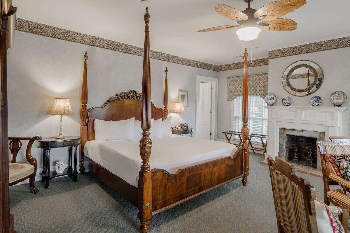 A four-poster bed with a wooden headboard, a ceiling fan, a fireplace, and a mirror on the wall.