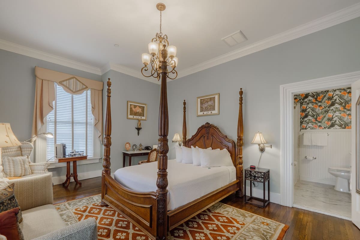 A four-poster bed with a wooden frame and white bedding, a chandelier, a patterned rug, and a bathroom with floral wallpaper.