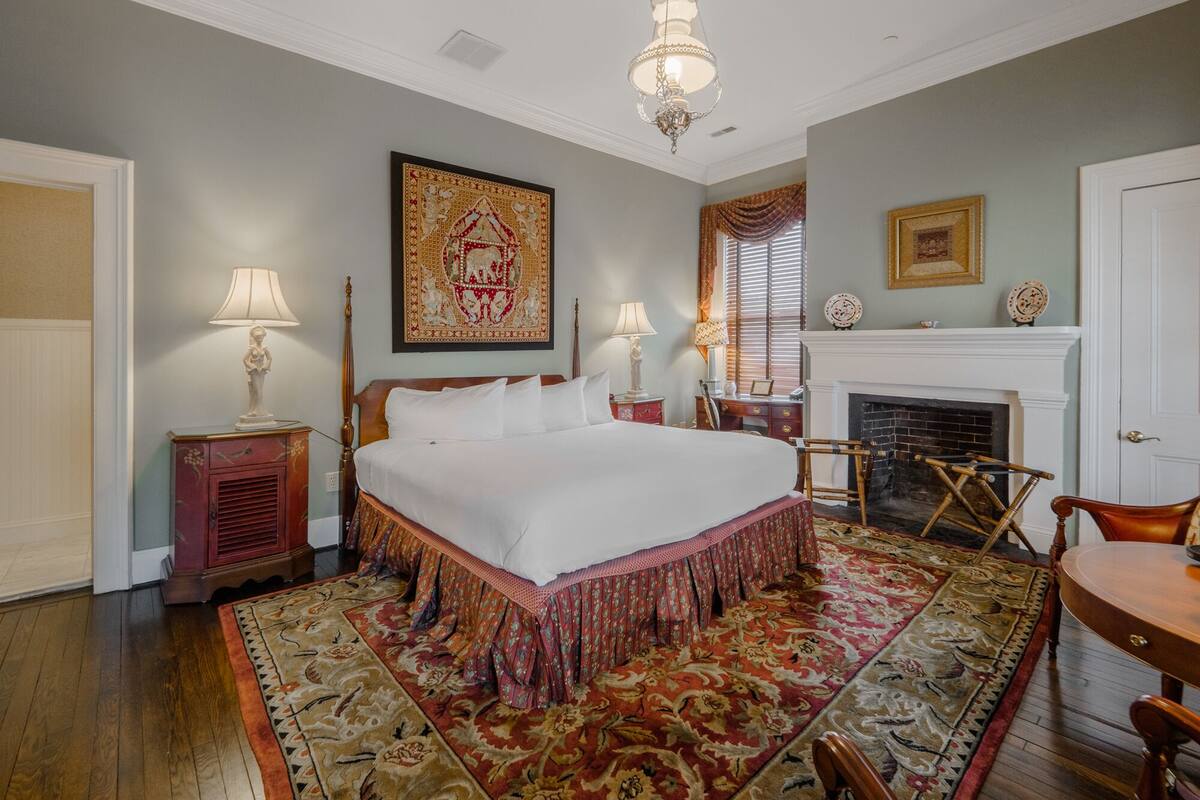 A bedroom with a large bed, two bedside tables with lamps, a fireplace, and a patterned rug.