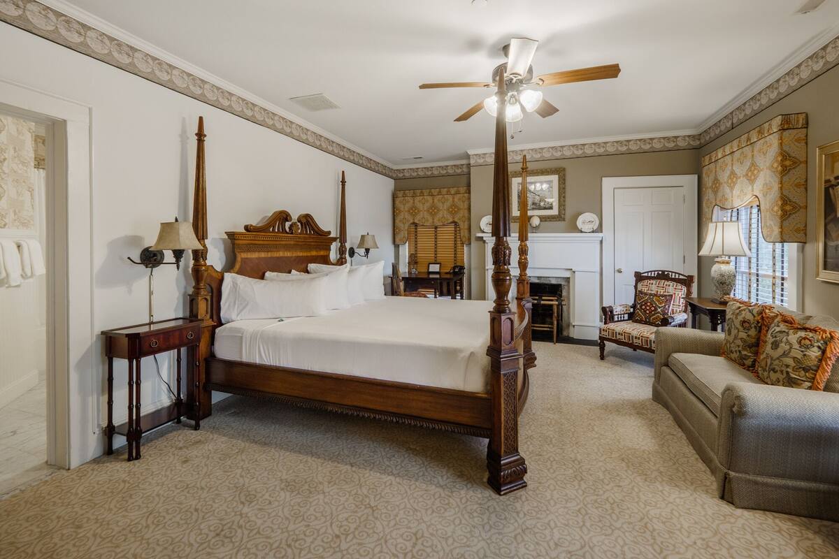 A four-poster bed with a ceiling fan, a fireplace, and a sofa in a room with patterned wallpaper and a carpet.