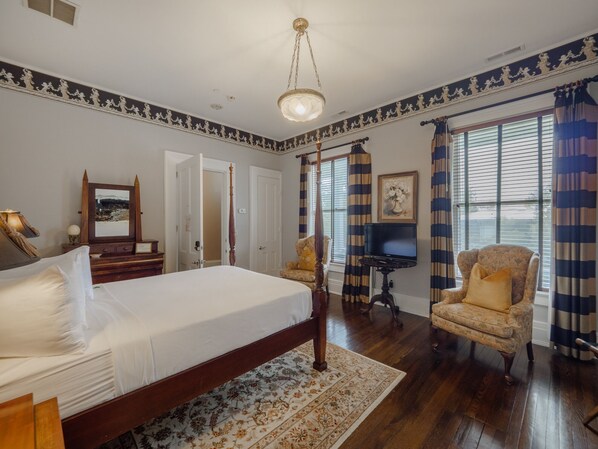 A bedroom with a four-poster bed, a chair, a television, and a large window with curtains. - The Verandas (Wilmington)