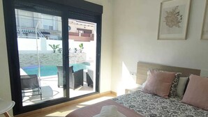 A hotel room with a bed, a large window with a view of a pool and buildings, and a chair by the window. - Independent villa with private pool, 5 min from the beach! (San pedro del Pinatar)