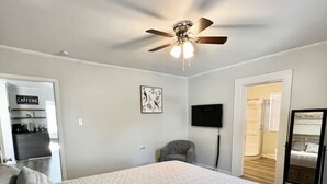 A bedroom with a large bed, a ceiling fan, a flat-screen TV, and a door leading to another room.