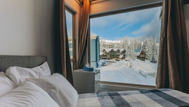 A bedroom with a large window, a bed, a bedside table, and a view of a snowy landscape with buildings. - The Fjäll | Ski-In Ski-Out Village Studio (Big White)
