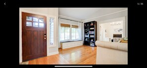 A spacious living room with a wooden floor, a large window with a view, a bookshelf, and a beige sofa.