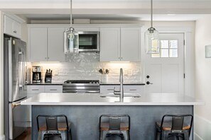 Cocina moderna con armarios blancos, electrodomésticos de acero inoxidable, una isla central con taburetes de bar e iluminación colgante.