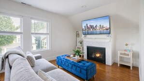 A living room with a gray sofa, a blue ottoman, a fireplace, and a TV mounted above it. - Stunning Home Mins to Boston w/Parking & Laundry (Somerville)