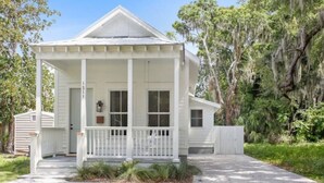 Une maison blanche, de plain-pied, dotée d’un porche et entourée d’une végétation luxuriante et d’arbres.