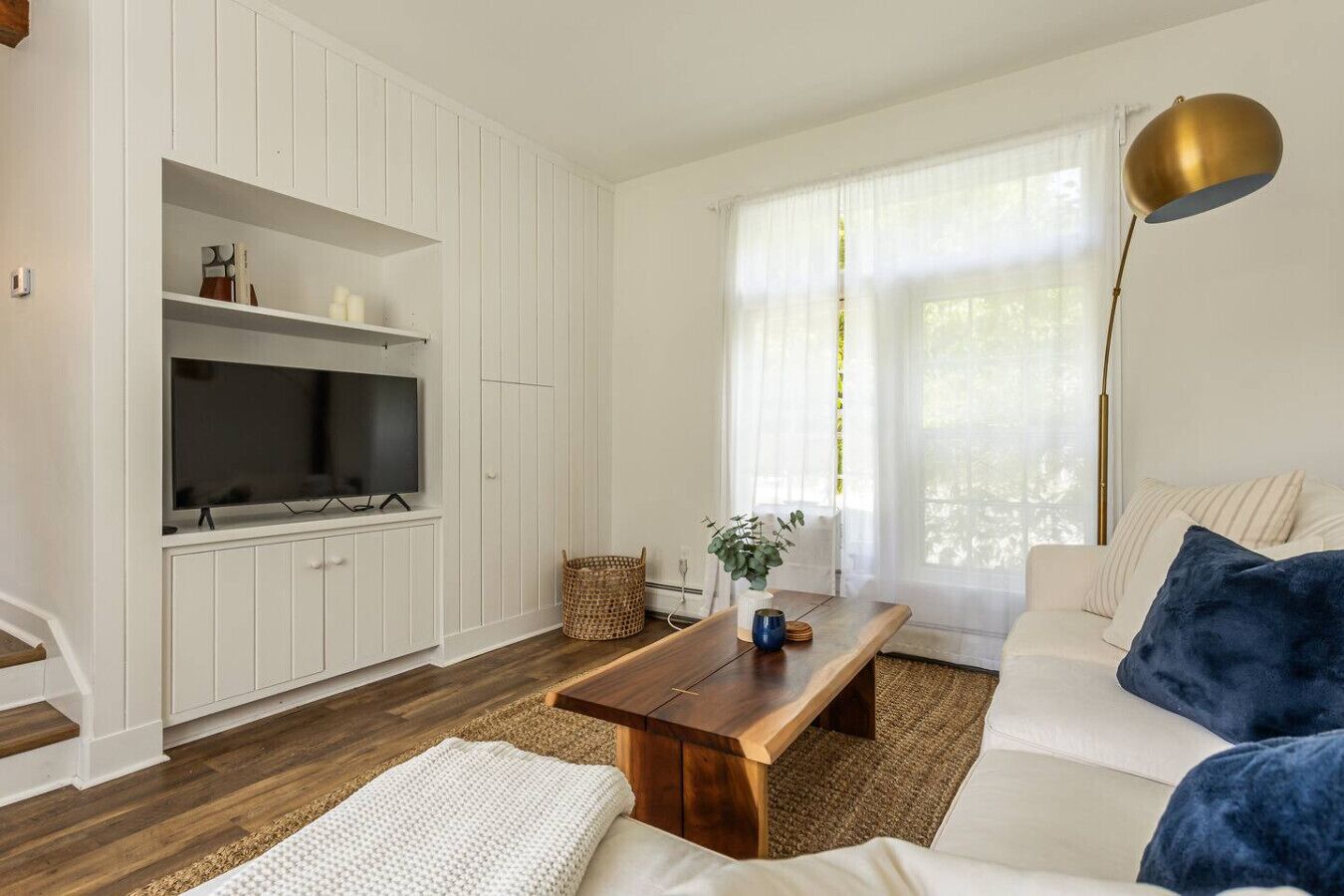 A modern living room with a television, wooden coffee table, white sofa, and a large window with sheer curtains.