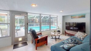 A spacious living room with a large window overlooking a swimming pool, a comfortable sofa, and a flat-screen TV.
