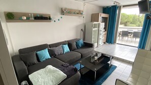 A modern living room with a grey sofa, a coffee table, and a ceiling fan. There is a kitchen area with a refrigerator and a dining table with chairs.