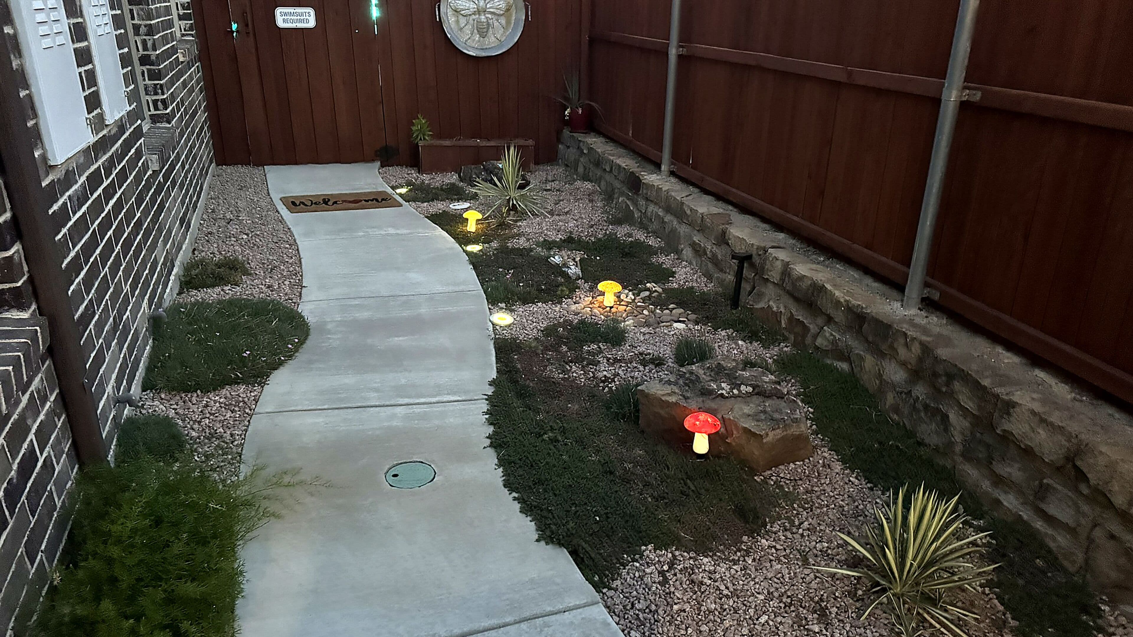 Ein Weg führt zu einem Holztor mit Schild, das Badebekleidungspflicht anzeigt, umgeben von einem angelegten Garten mit Steinen, Pflanzen und einer Willkommensmatte.